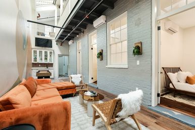 Bright modern industrial loft living room with skylight and mezzanine, orange velvet sectional, woven wooden chairs with faux-fur throws, exposed painted brick wall, kitchenette and open bedroom