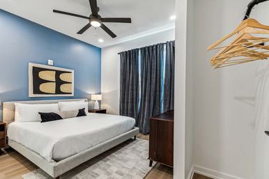 Contemporary apartment bedroom with king-size bed, blue accent wall and abstract art, black ceiling fan, bedside lamps, dark floor-to-ceiling curtains, wooden dresser and open closet with wooden hangers