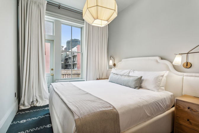 Bright modern bedroom with a large white bed, upholstered headboard, gray throw and lumbar pillow, wall sconces and wooden nightstand; tall window and glass door open to an urban city balcony view.