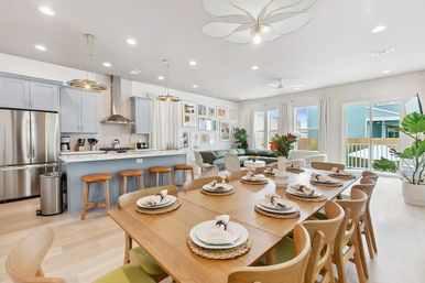 Bright, airy open-plan kitchen and living room with pale-blue island, stainless appliances, wooden bar stools, long wooden dining table set for ten, cozy green sofa, gallery wall and sliding glass doors letting in natural light.