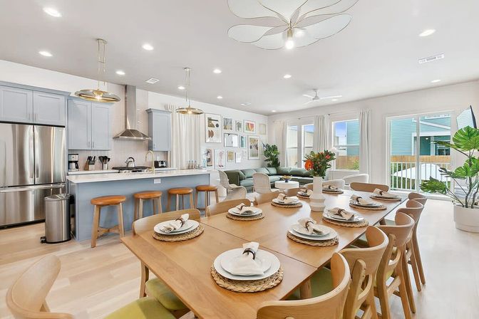 Bright, airy open-plan kitchen and living room with pale-blue island, stainless appliances, wooden bar stools, long wooden dining table set for ten, cozy green sofa, gallery wall and sliding glass doors letting in natural light.