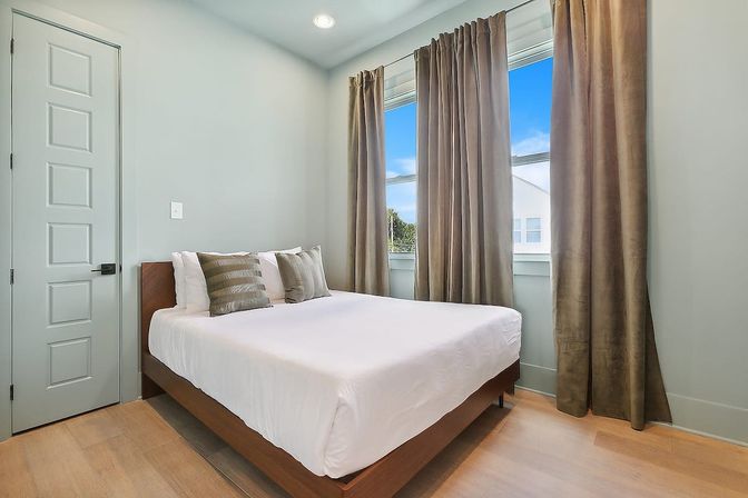 Sunlit modern urban apartment bedroom with queen bed, white linens, wood platform frame, large window with taupe curtains and light hardwood floors.