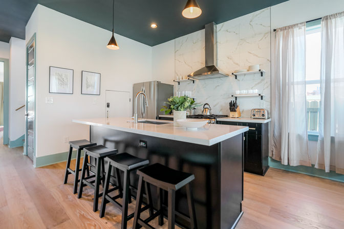Bright modern open kitchen with a black island and four bar stools, white quartz countertop, stainless-steel fridge and range, marble-effect backsplash, pendant lights, hardwood floors and a sunlit window.
