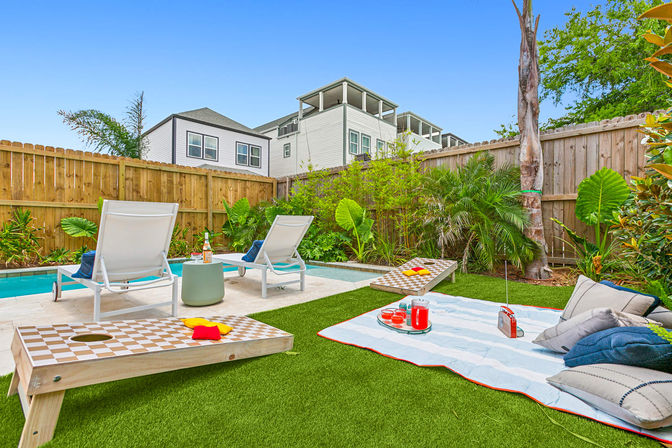 Sunny backyard oasis with a small pool, two white lounge chairs, cornhole boards and a striped picnic blanket with cushions, drinks and a radio on artificial turf, fenced garden and modern homes beyond