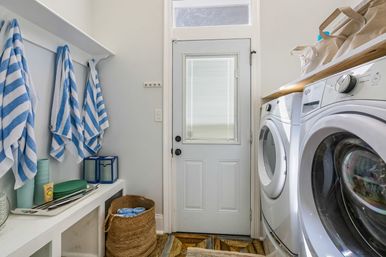 Bright entry laundry room with side-by-side white front‑load washer and dryer under a wood countertop, white door with window, blue-and-white striped towels on hooks, woven laundry basket and cubby storage.