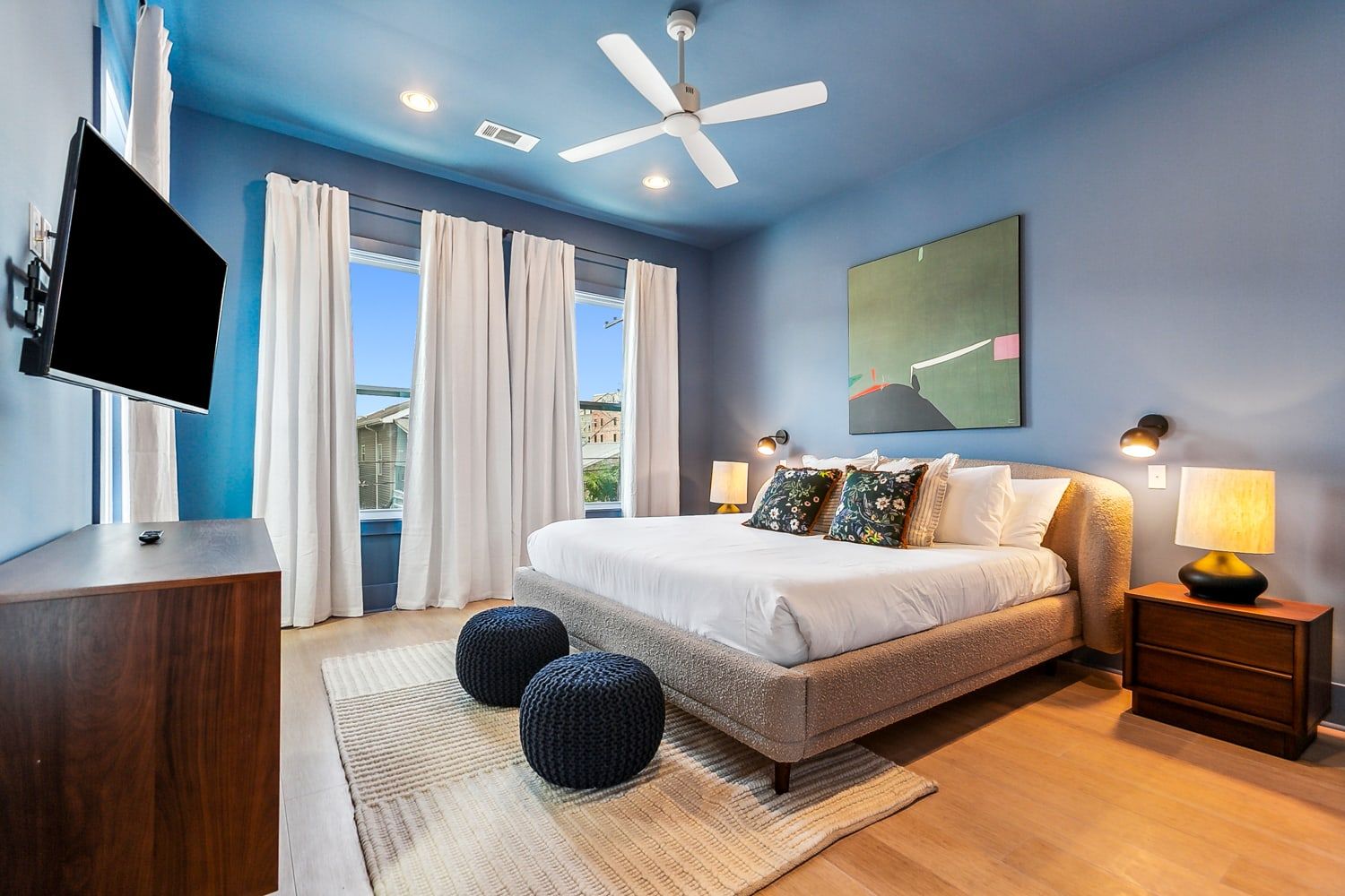Stylish sunlit blue bedroom with king bed and white linens, patterned throw pillows, two navy knit poufs, wood nightstands with lamps, wall-mounted TV, ceiling fan and large windows with white curtains.
