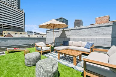 Sunny urban rooftop lounge with modern wooden-framed sofas, wooden coffee table, beige umbrella, patterned poufs and a cornhole board on artificial turf with city buildings in the background.