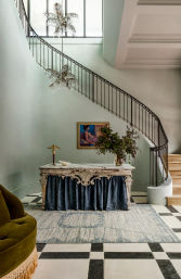 Elegant foyer with a sweeping curved staircase and wrought-iron railing, black-and-white checkered tile floor, ornate marble console draped in blue fabric with a vase of greenery, crystal palm chandeliers and framed artwork on pale green walls.