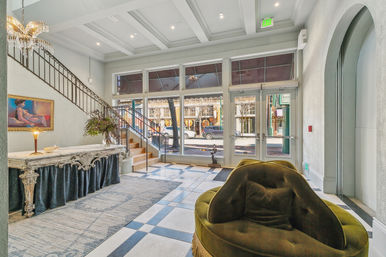 Bright hotel lobby with coffered ceiling and crystal chandelier, ornate marble console table with floral arrangement, curved iron staircase, floor-to-ceiling windows and glass doors facing a downtown street, tiled checkerboard floor and plush olive-green velvet lounge chair.