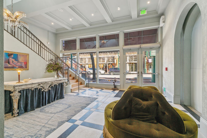 Bright hotel lobby with coffered ceiling and crystal chandelier, ornate marble console table with floral arrangement, curved iron staircase, floor-to-ceiling windows and glass doors facing a downtown street, tiled checkerboard floor and plush olive-green velvet lounge chair.