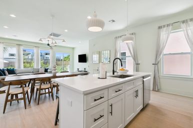 Bright modern open-concept kitchen and dining area in a suburban home with a white island and black faucet, pendant lights, wood dining table and chairs, large windows with sheer curtains, and light hardwood floors.