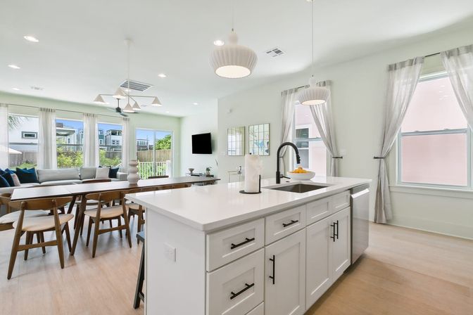 Bright modern open-concept kitchen and dining area in a suburban home with a white island and black faucet, pendant lights, wood dining table and chairs, large windows with sheer curtains, and light hardwood floors.