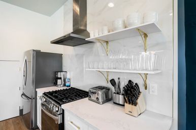 Bright modern white kitchen corner with stainless steel fridge and gas range, marble-look backsplash, gold-bracket open shelves with glassware, toaster, coffee maker, and wooden knife block.