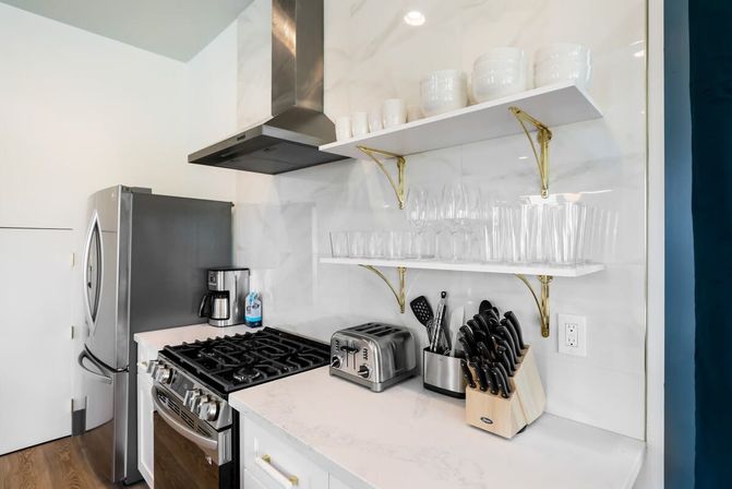 Bright modern white kitchen corner with stainless steel fridge and gas range, marble-look backsplash, gold-bracket open shelves with glassware, toaster, coffee maker, and wooden knife block.