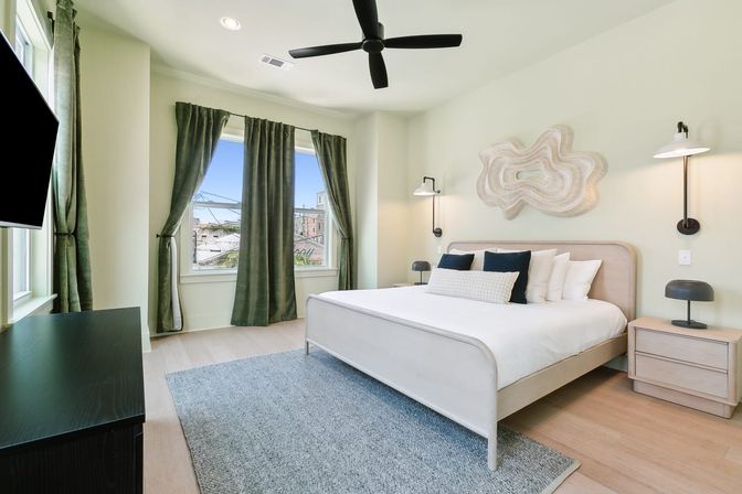 Bright modern master bedroom with king bed, white bedding and navy pillows, abstract wall art, black ceiling fan, matching nightstands and lamps, green floor-length curtains framing large windows with urban rooftop views.