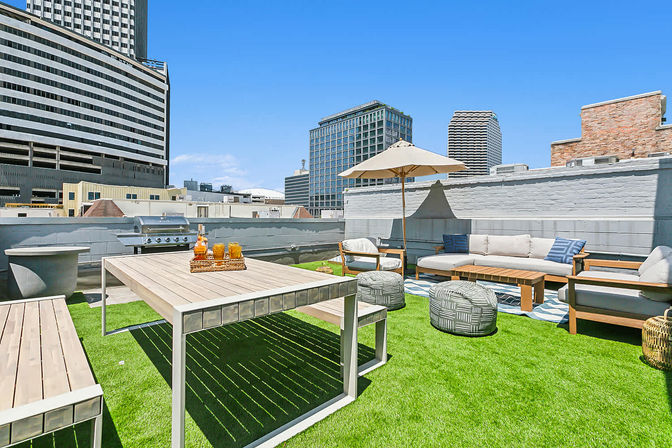 Sunny urban rooftop patio with artificial turf, modern dining table, cushioned sofa and chairs, umbrella and poufs, framed by downtown skyscrapers