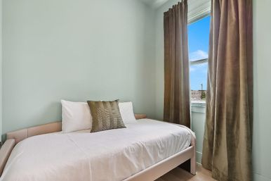 Cozy light-filled apartment bedroom with a single white bed, pale green wall, textured accent pillow and floor-length brown velvet curtains framing a window with blue sky and rooftop view.