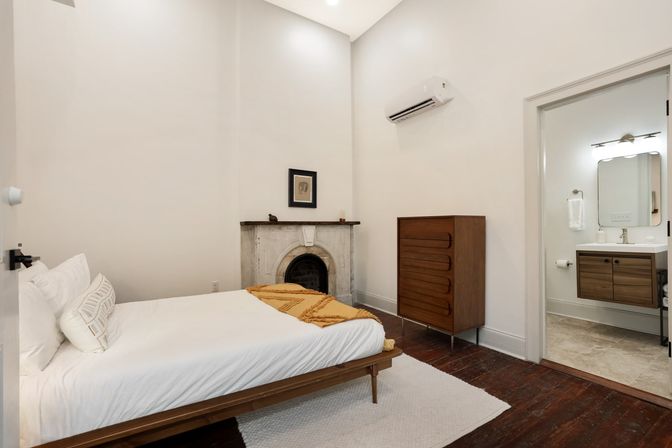 Cozy minimalist bedroom with high white walls and dark hardwood floors, low wooden platform bed with white linens and mustard throw, small marble fireplace, mid-century wooden dresser, wall-mounted air conditioner, and open door to a modern ensuite bathroom.