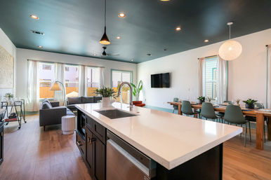 Modern open-concept kitchen and living area featuring a large white quartz island with sink, dark painted ceiling with pendant lights, wood floors, gray sofa by sheer-windowed wall, mounted TV, long wooden dining table with green chairs and potted plants.