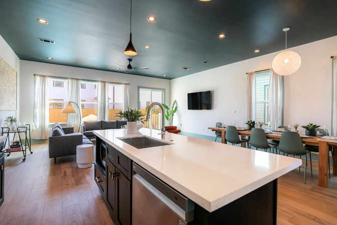 Modern open-concept kitchen and living area featuring a large white quartz island with sink, dark painted ceiling with pendant lights, wood floors, gray sofa by sheer-windowed wall, mounted TV, long wooden dining table with green chairs and potted plants.
