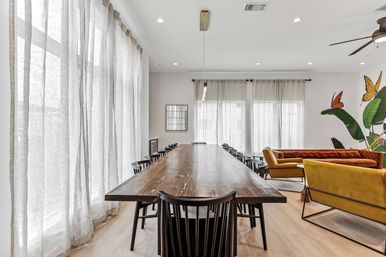 Bright modern communal lounge with a long dark wooden table surrounded by black chairs, floor-to-ceiling sheer curtains, mustard and rust velvet sofas, a tropical leaf and butterfly wall mural, and light wood floors.