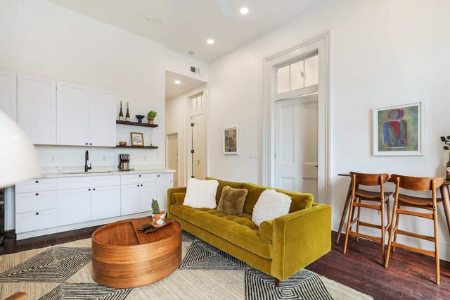 Modern open-concept apartment living room with olive-green velvet sofa, white kitchenette with floating shelves, round wooden coffee table on a patterned rug, and wooden bar stools under high ceilings.