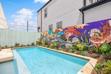Backyard rectangular swimming pool beside a colorful fence mural of a blue‑haired mermaid playing a guitar with oversized fish and lush tropical landscaping