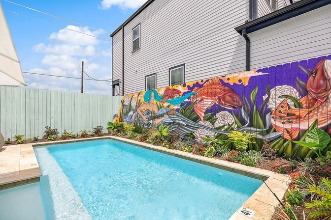 Backyard rectangular swimming pool beside a colorful fence mural of a blue‑haired mermaid playing a guitar with oversized fish and lush tropical landscaping