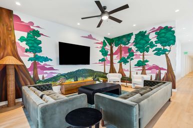 Bright modern communal lounge with velvet tufted sofas, white armchairs, wall-mounted TV and ceiling fan, light wood floors, and a playful wall mural of trees and a giant green crocodile.