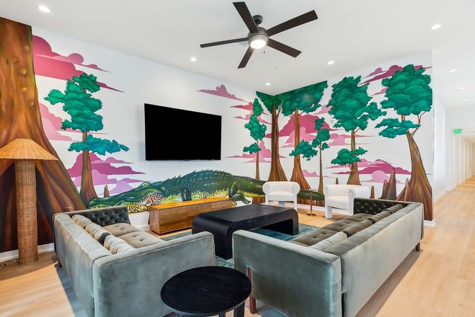Bright modern communal lounge with velvet tufted sofas, white armchairs, wall-mounted TV and ceiling fan, light wood floors, and a playful wall mural of trees and a giant green crocodile.