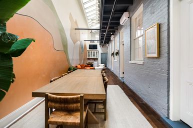 Long wooden communal dining table in a bright urban loft with a skylight, abstract mural on a peach wall, gray exposed-brick opposite wall, woven chairs and hardwood floors