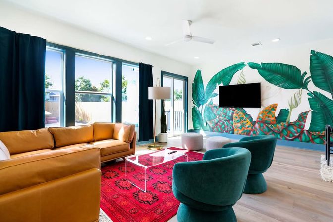 Bright modern living room with large windows and natural light, tan leather sectional, teal velvet swivel chairs, red patterned rug, clear acrylic coffee table, floor lamp, wall-mounted TV and a vibrant tropical leaf mural on white wall over hardwood floors.