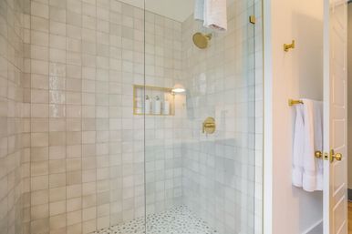 Bright modern glass-enclosed walk-in shower with white square wall tiles, brass showerhead and controls, recessed niche with bath bottles, pebble tile floor and white towels