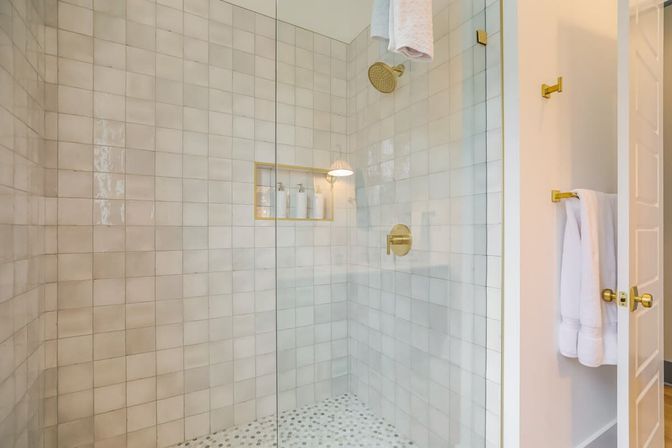 Bright modern glass-enclosed walk-in shower with white square wall tiles, brass showerhead and controls, recessed niche with bath bottles, pebble tile floor and white towels