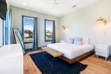 Sunlit modern bedroom with king bed in white linens, pastel pillows, gold wall sconces, tufted bench and navy rug; three large windows with blue curtains overlooking a neighborhood and distant water, hardwood floors and full-length mirror.