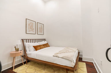 Cozy minimalist bedroom in a modern apartment: low wooden platform bed with white linens, rust velvet lumbar pillow, cream textured throw, pink round nightstand with lamp and brass animal figurine, neutral abstract art and yellow rug.