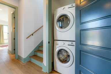 Stacked white front-loading washer and dryer in a compact hallway laundry nook beside a blue paneled door, light oak floors and mint-green trimmed staircase
