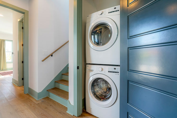 Stacked white front-loading washer and dryer in a compact hallway laundry nook beside a blue paneled door, light oak floors and mint-green trimmed staircase