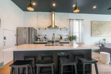 Bright modern open-concept kitchen with marble backsplash, stainless steel appliances, central island with white countertop and four black bar stools, pendant lights and open shelving.