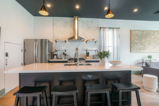Bright modern open-concept kitchen with marble backsplash, stainless steel appliances, central island with white countertop and four black bar stools, pendant lights and open shelving.