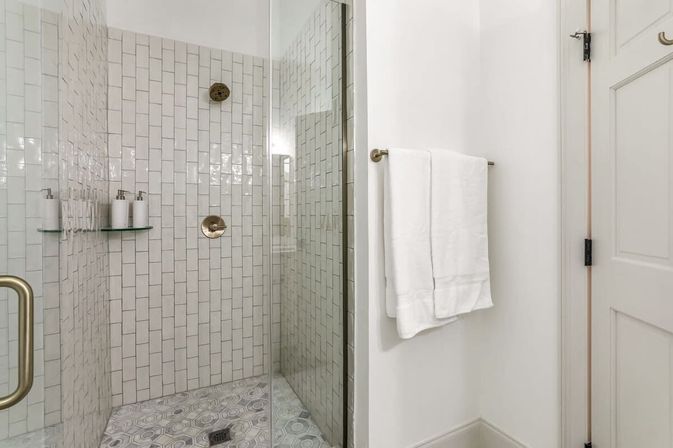 Bright spa-like walk-in bathroom shower with glass door, vertical white subway tiles, brass fixtures, hex-pattern floor tile, corner glass shelf with soap dispensers, and a folded white towel on a wall bar.