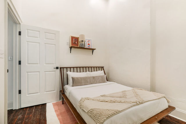 Cozy minimalist city apartment bedroom with a wooden mid-century bedframe, white linens and beige throw, small wall shelf holding a lamp and framed art, white paneled door and dark hardwood floors