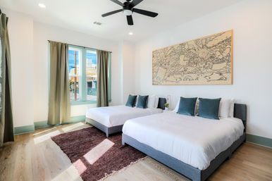 Bright, airy modern bedroom with two queen beds in gray frames, teal throw pillows, large doodle-style wall art, black ceiling fan, hardwood floors with a brown area rug, and a sunlit green-trimmed window with curtains.