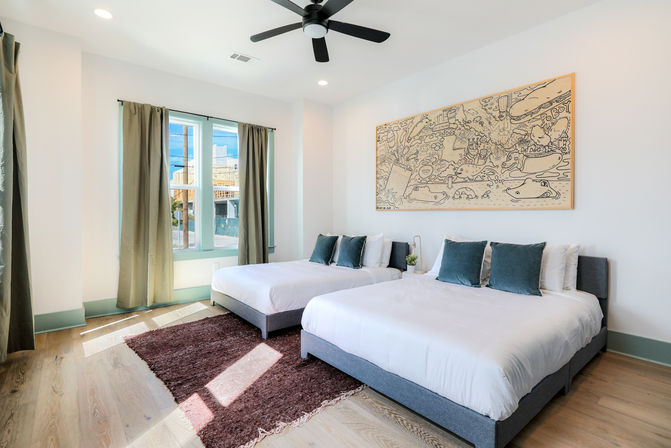 Bright, airy modern bedroom with two queen beds in gray frames, teal throw pillows, large doodle-style wall art, black ceiling fan, hardwood floors with a brown area rug, and a sunlit green-trimmed window with curtains.