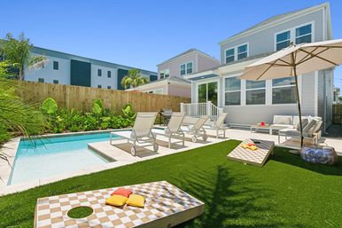 Sunny suburban backyard with a rectangular plunge pool, tiled patio and four white chaise lounges, umbrella-shaded outdoor seating, artificial turf with cornhole boards, palm landscaping and a modern two-story house.