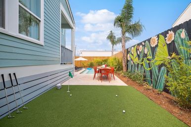 Coastal-style backyard with artificial putting green and golf clubs in the foreground, concrete patio with red dining set, small swimming pool, palm trees, and a vibrant floral mural on a wooden fence under a blue sky.