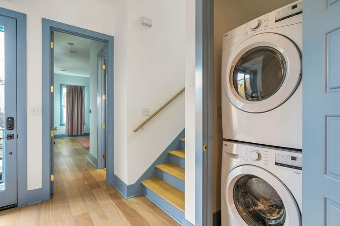 Sunlit entry hall with light hardwood floors, blue-painted trim and staircase, and a stacked white washer and dryer tucked into a closet