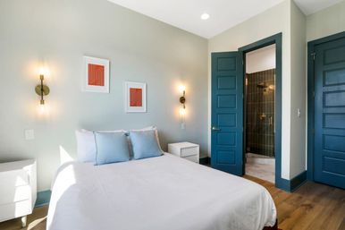Bright modern bedroom with white bedding and light blue pillows, teal paneled doors opening to a tiled ensuite shower, brass wall sconces, wood floor and minimalist nightstands.