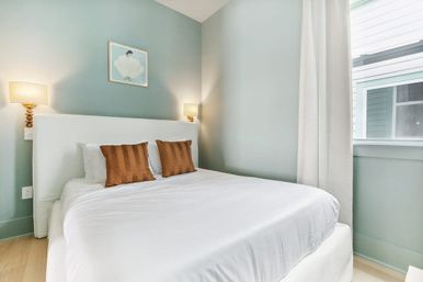 Bright, cozy bedroom with white bed and linens, two rust accent pillows, seafoam-green walls, wall sconces, and a sunlit window