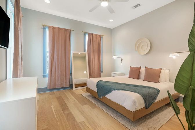 Minimal modern bedroom interior with low wooden platform bed, neutral walls, pink curtains, light oak floors, full-length mirror, green throw and potted plant.